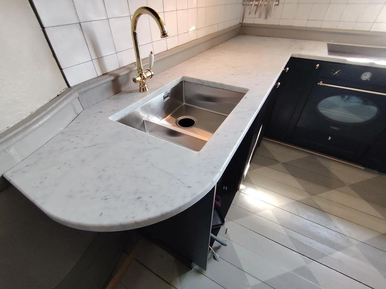 Kurvad köksbänk i matt Carrara C marmor med underlimmad rostfri diskho och mässingsfärgad blandare i ett kök i Örsundsbro.