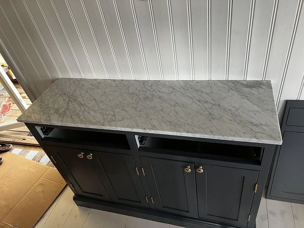 Köksbänkskiva i vit Carrara C marmor med grå ådring, matt yta, på marinblå köksstommar med mässingsbeslag i ett kök i Prässebo.