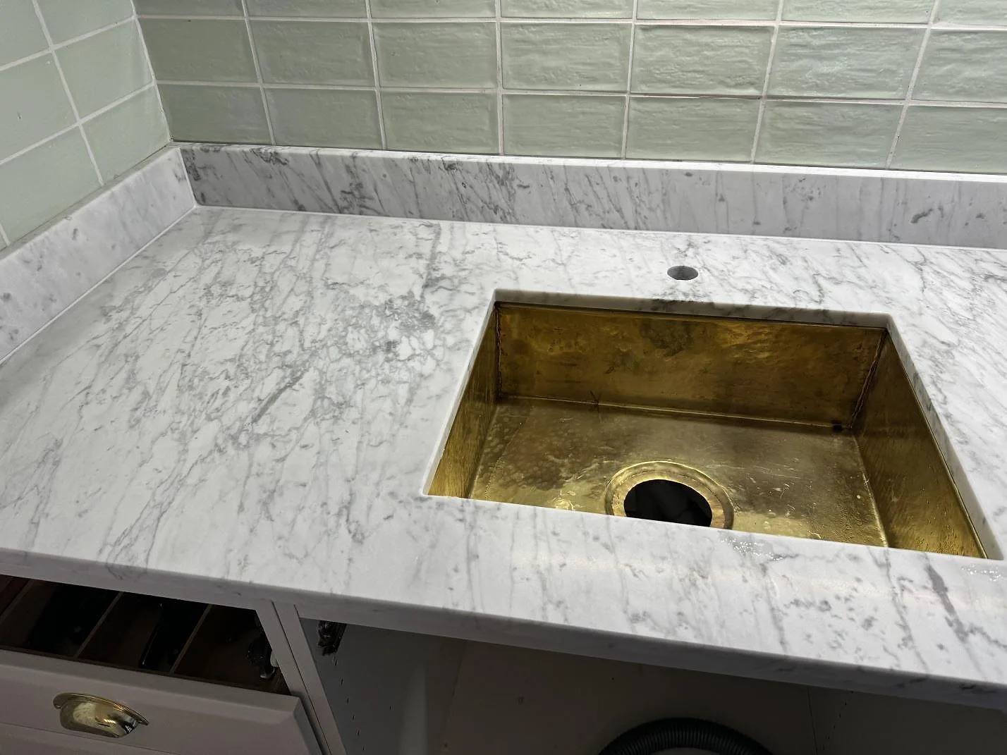 Carrara C marmorbänkskiva i ett kök i Rockneby, med mässingsfärgad diskho under stenen och gröna väggplattor bakom.