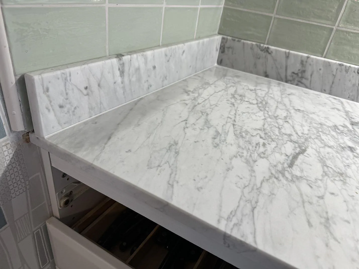 Närbild på matt Carrara C marmor som köksbänkskiva med uppvikt stänkkant och grön kakelvägg i Rockneby.