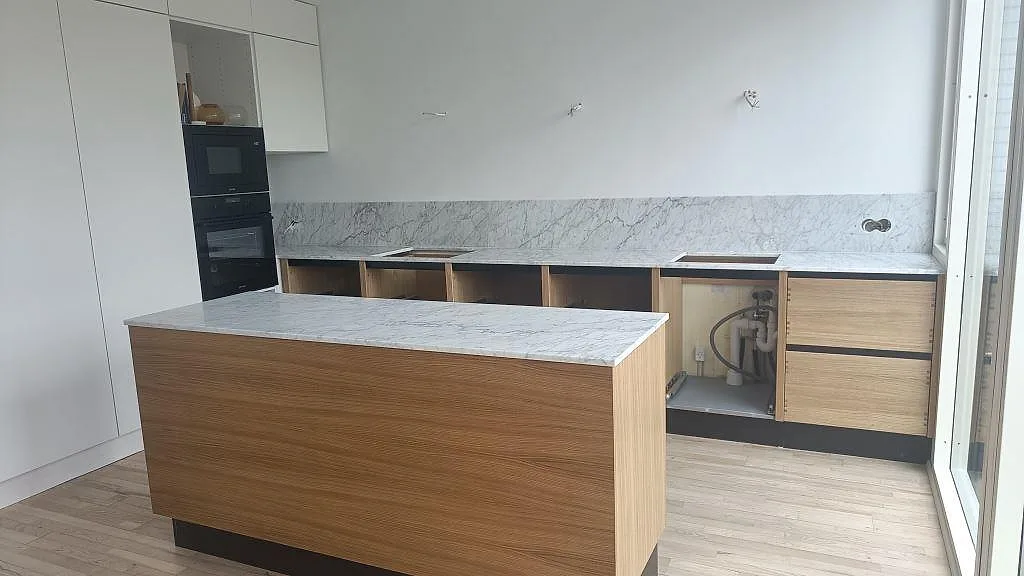 Köksbänkskiva och köksö i vit Carrara C-marmor med grå ådring, monterad på ekstommar med stänkskydd i samma marmor, i ett kök i Rødovre.