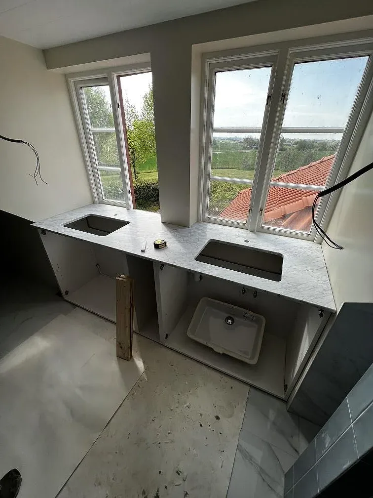 Badrumsbänkskiva i matt Carrara C marmor, 20 mm, med vitt tvättställ och väggbeklädnad under fönster i Rydsgård.