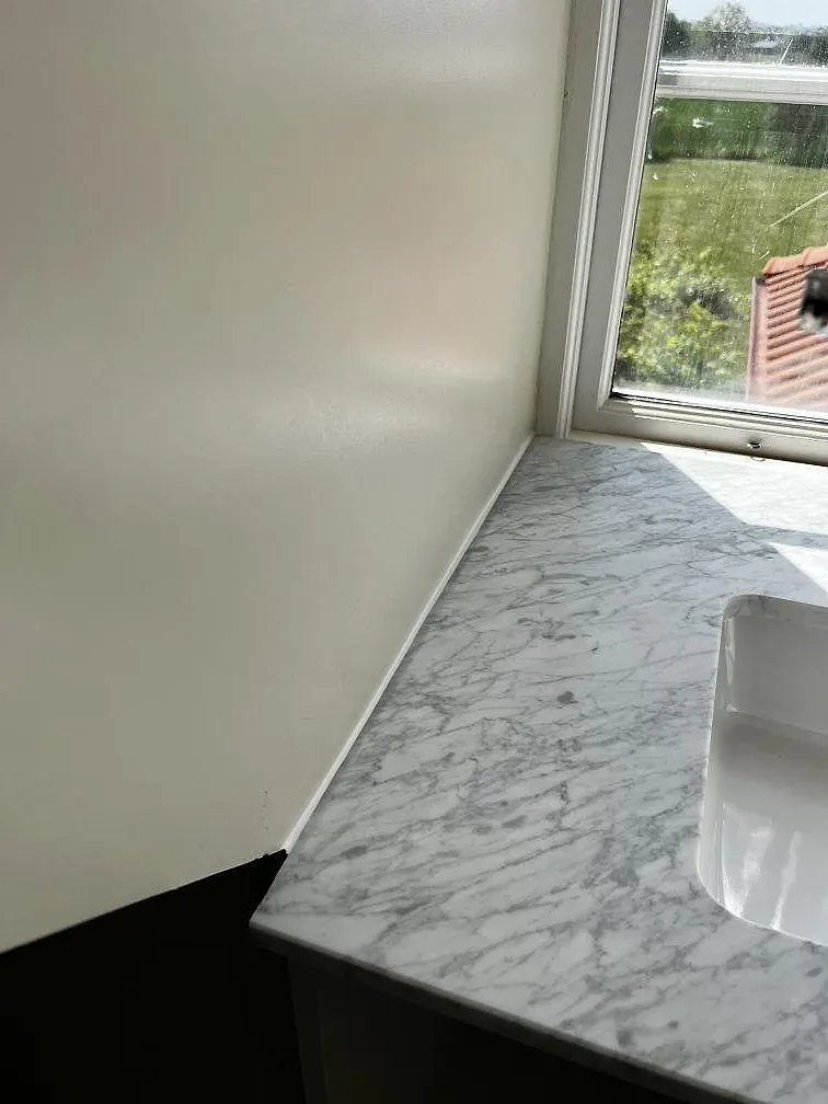 Detalj av frontkant och väggbeklädnad i matt Carrara C marmor vid ett badrumstvättställ i Rydsgård.