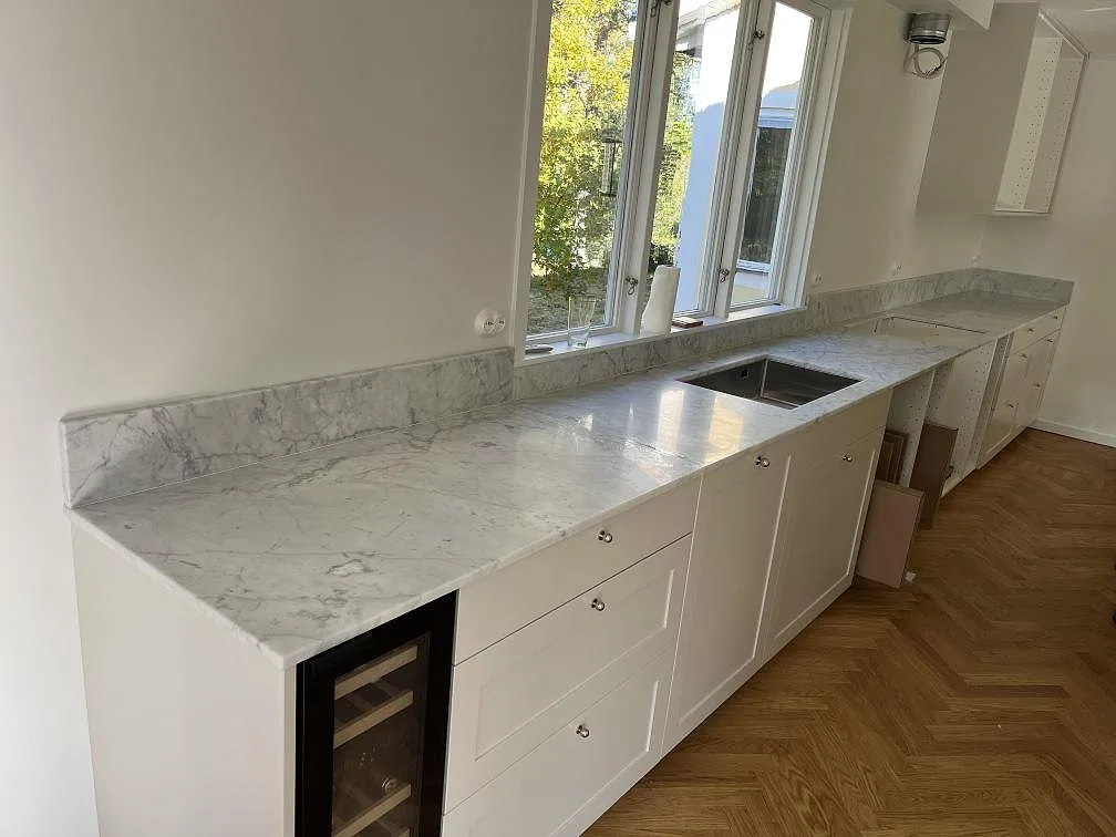 Köksbänkskiva i matt Carrara C marmor i ett L-format kök i Saltsjö Duvnäs, med låg stänkkant, underlimmad dubbel rostfri diskho och vinkyl.