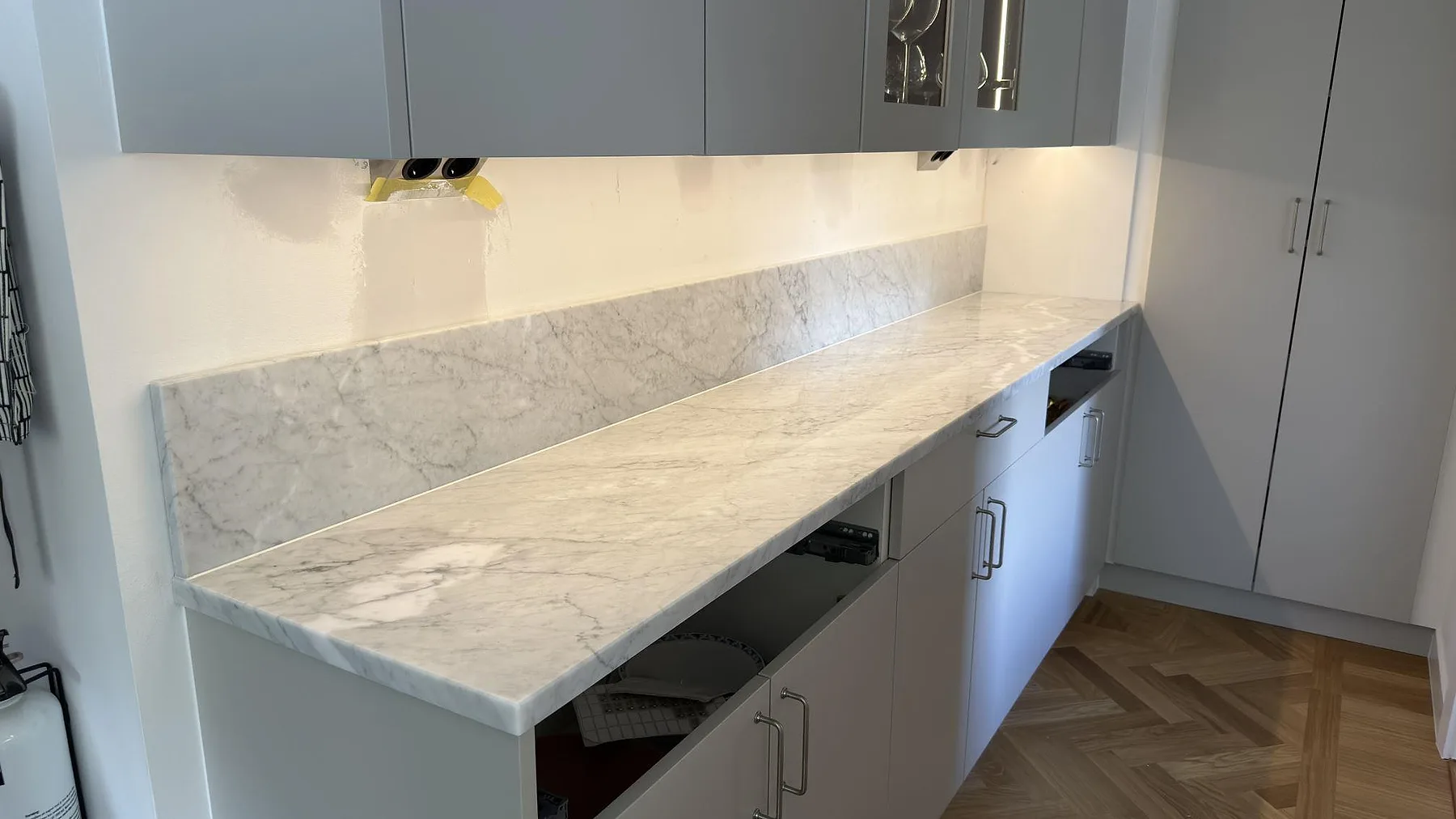 Lång köksbänkskiva i polerad Carrara C marmor med sammanhängande stänkkant och öppna stommar under montering i Saltsjöbaden.