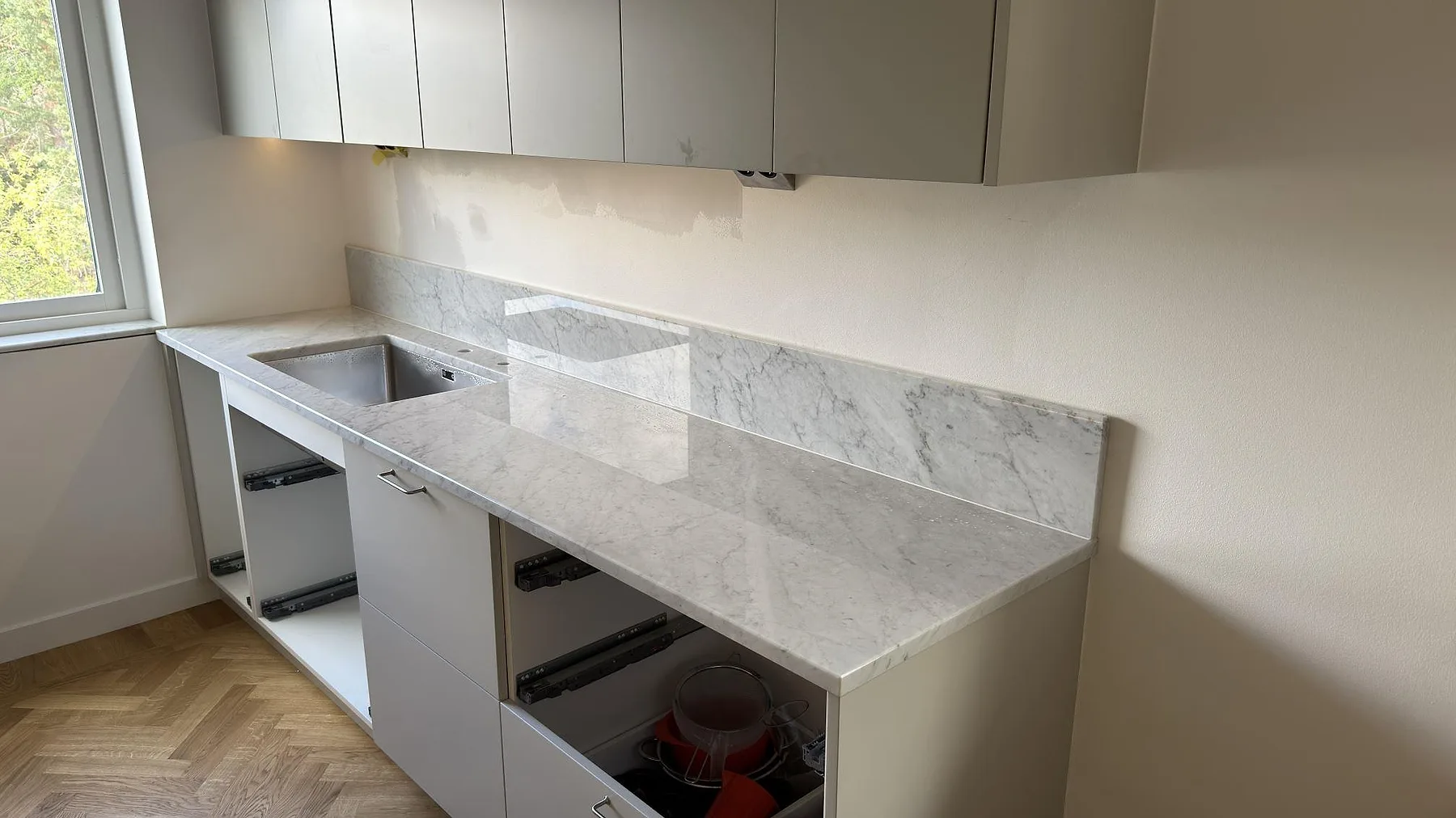 Köksbänkskiva i polerad Carrara C marmor med låg stänkkant och diskhozon i ett kök under montering i Saltsjöbaden.
