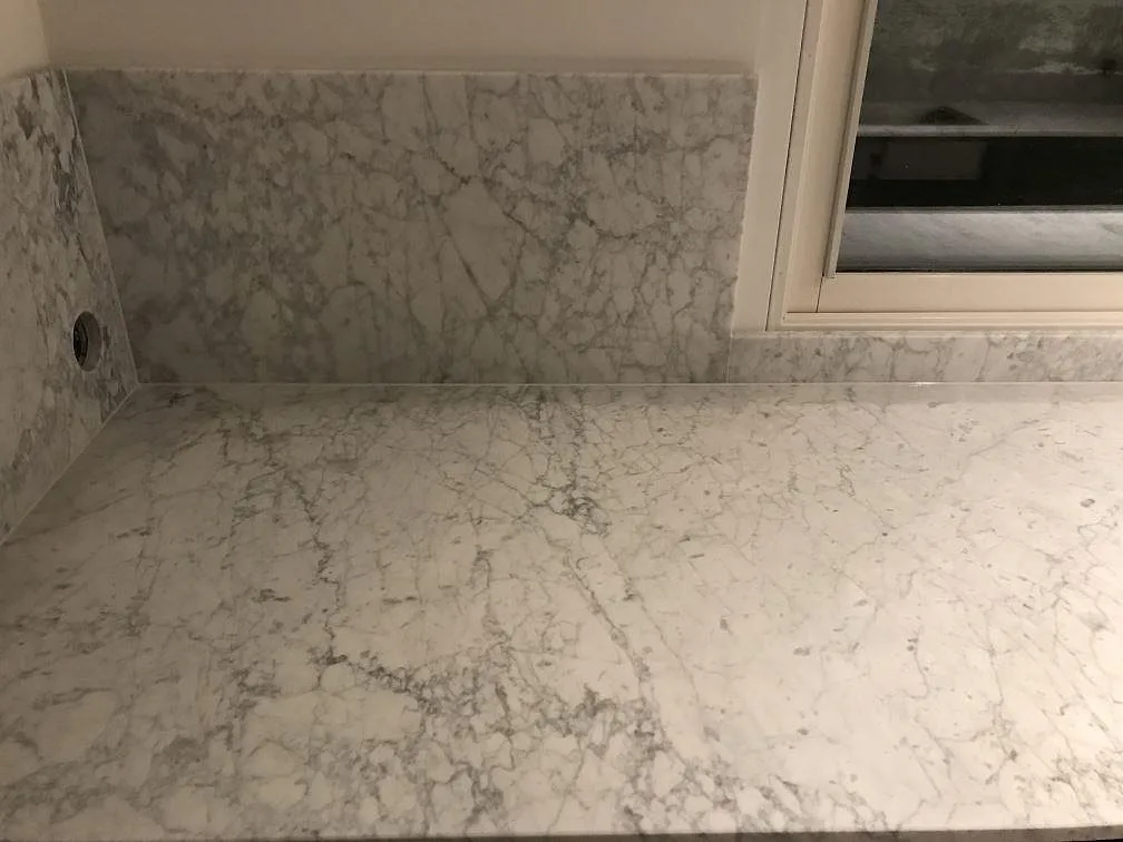 Köksbänkskiva i vit Carrara C marmor, matt, med fönsterbänk och uppvikt stänkskydd i samma sten vid fönstret i ett kök i Sandared.