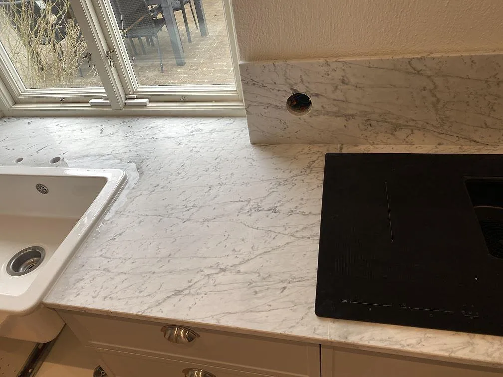 Köksbänkskiva i vit Carrara C marmor med vit keramikho, induktionshäll och uppvikt stänkkant vid fönstret i ett kök under montering i Skanör.