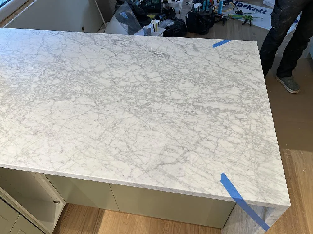 Närbild av matt Carrara C marmor på köksö med vattenfallskant och blå monteringstejp i ett kök i Skanör.