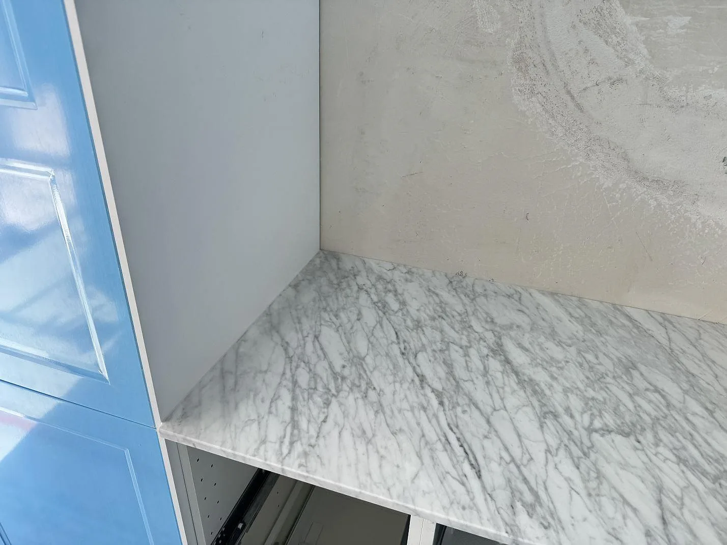 Närbild av polerad Carrara C-marmorbänkskiva mot rå vägg och blå köksfront under montering i Skillinge.