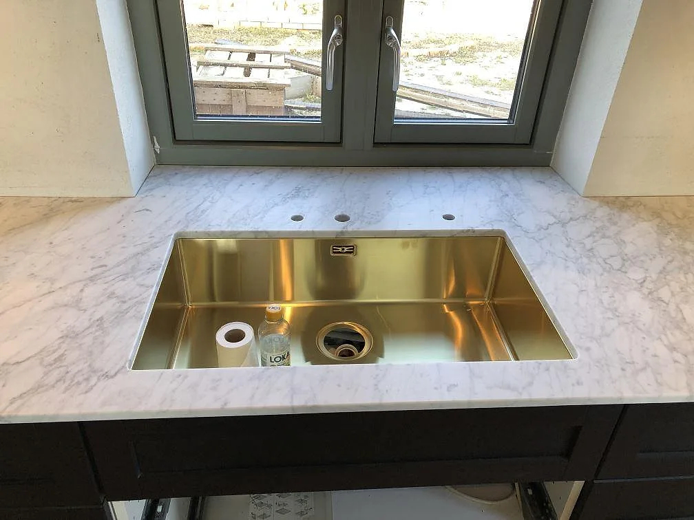 Underlimmad mässingsfärgad diskho i en bänkskiva av Carrara C-marmor under fönstret i ett kök i Skurup.