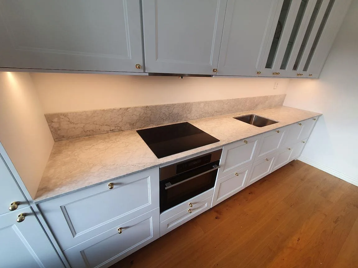 Köksbänk i matt Carrara C marmor med induktionshäll och rostfri diskho i ett kök i Stockholm, sedd i vinkel med stänkkanten i bakgrunden.