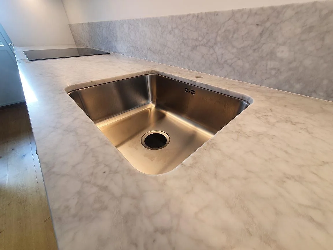 Närbild på underlimmad rostfri diskho i en bänkskiva av matt Carrara C marmor med levande ådring i ett kök i Stockholm.