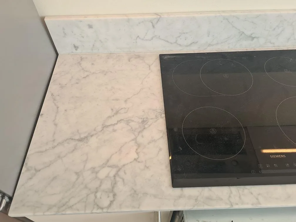 Närbild av matt Carrara C marmor runt svart induktionshäll och marmorstänkkant i kök i Stockholm.