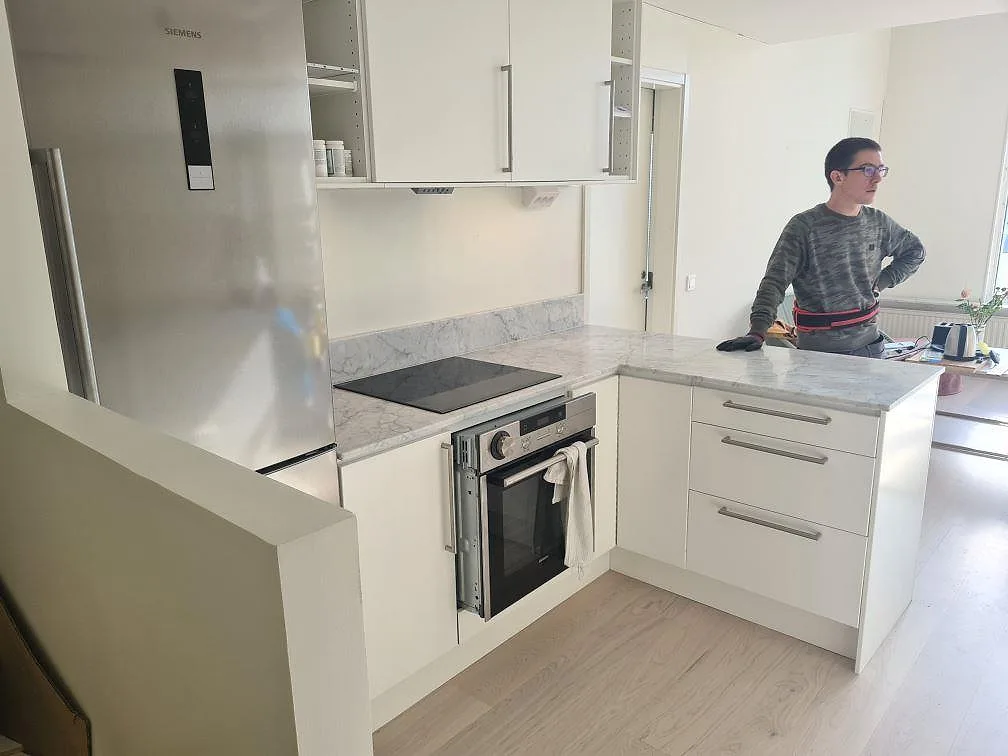 Köksbänkskiva i matt Carrara C marmor med uppvikt stänkkant, svart induktionshäll och vit köksinredning i Stockholm.