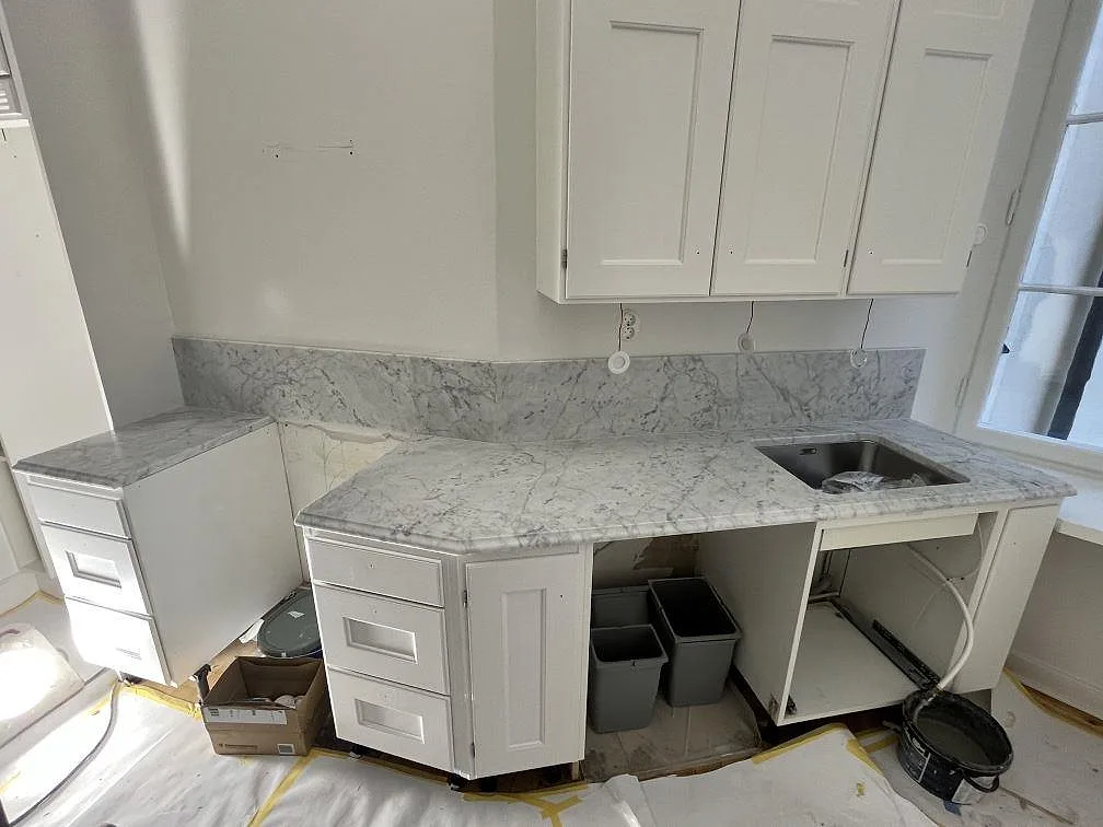L-formad köksbänkskiva i Carrara C marmor, matt, med uppvikt stänkskydd och underlimmad diskho i ett vitt kök i Stockholm under montering.