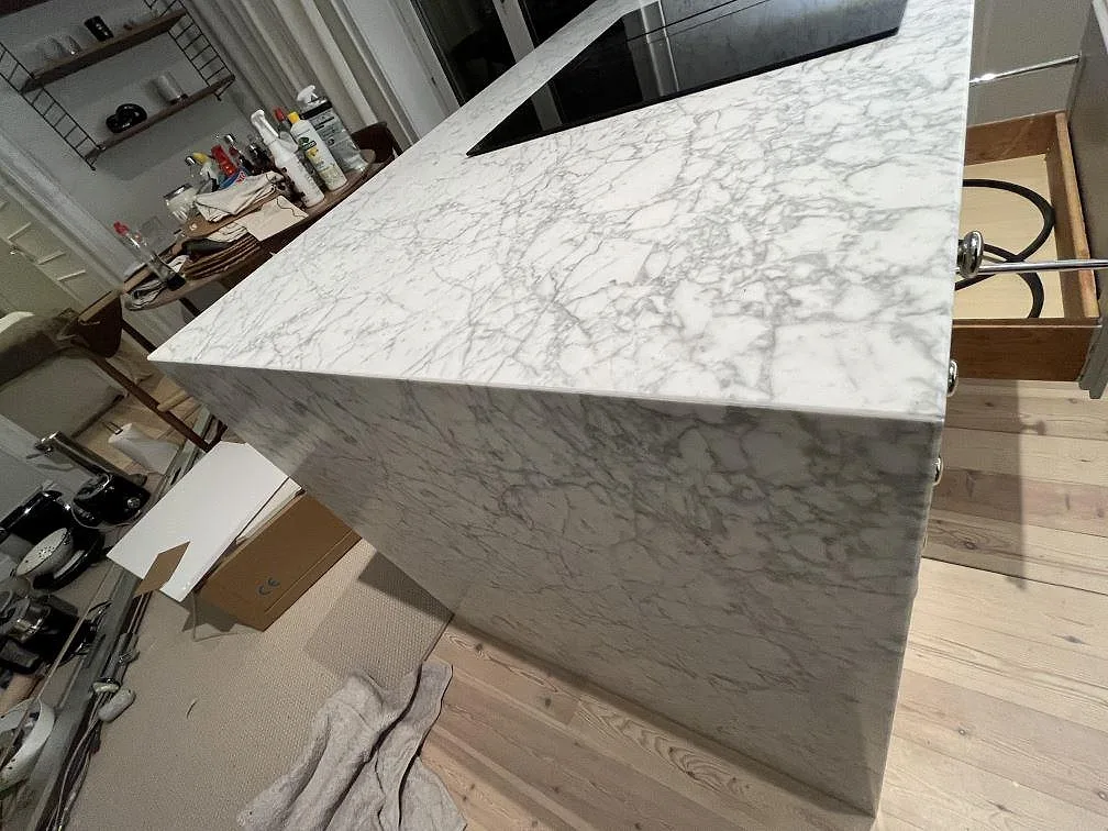 Detalj av kökshalvö i matt Carrara C-marmor i Stockholm, med svart induktionshäll, synlig ådring och neddragen stengavel.