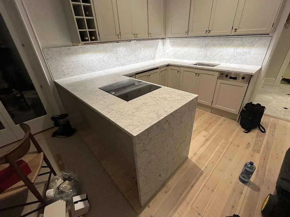 Köksbänkskiva i matt Carrara C-marmor i ett U-format kök i Stockholm, med neddragen waterfall-gavel, svart induktionshäll och ljus sten mot vita skåp.