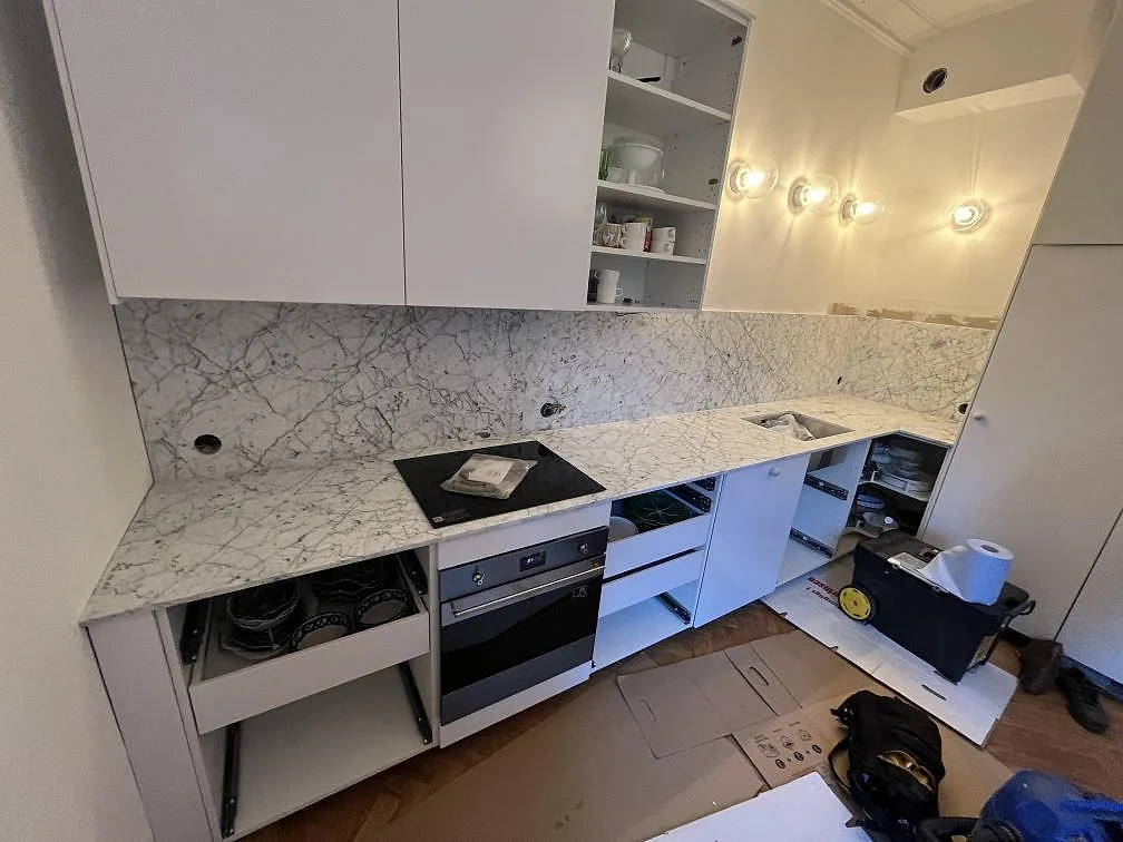 Köksbänkskiva och stänkskydd i matt Carrara C marmor med synlig häll och ugn i ett vitt kök i Stockholm under montering.