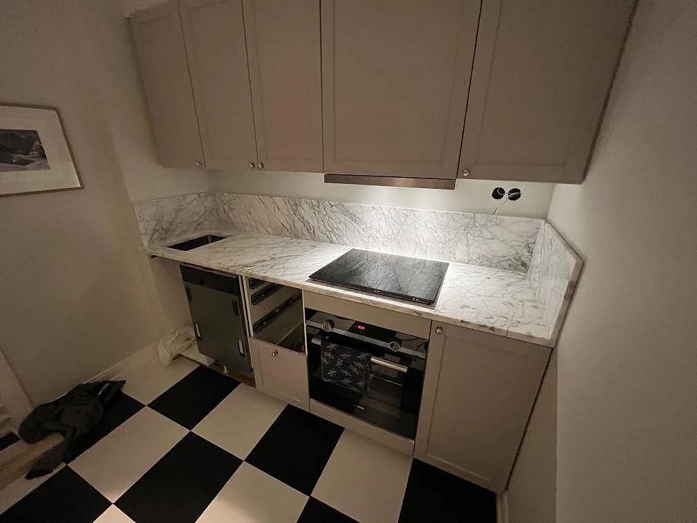 Polerad köksbänkskiva i Carrara C marmor med stänkskydd, häll och schackrutigt golv i ett kök i Stockholm.