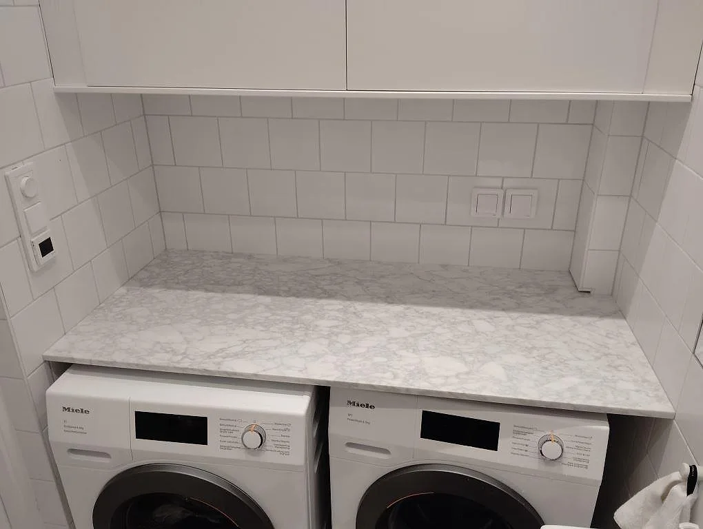 Bänkskiva i polerad Carrara C marmor, 20 mm, monterad som tvättstugeyta över två tvättmaskiner i Stockholm.