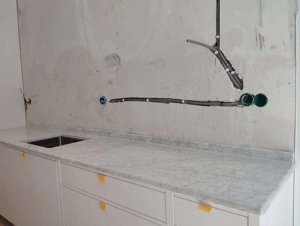 Köksbänkskiva i polerad Carrara C marmor med underlimmad rostfri diskho, uppvikt stänkkant och råputsad vägg i Stockholm.