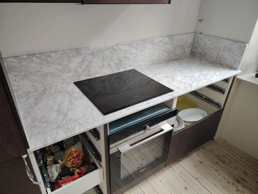 Väggbänk i matt Carrara C marmor med svart induktionshäll, låg stenkant och mörka köksstommar i ett kök i Stockholm.