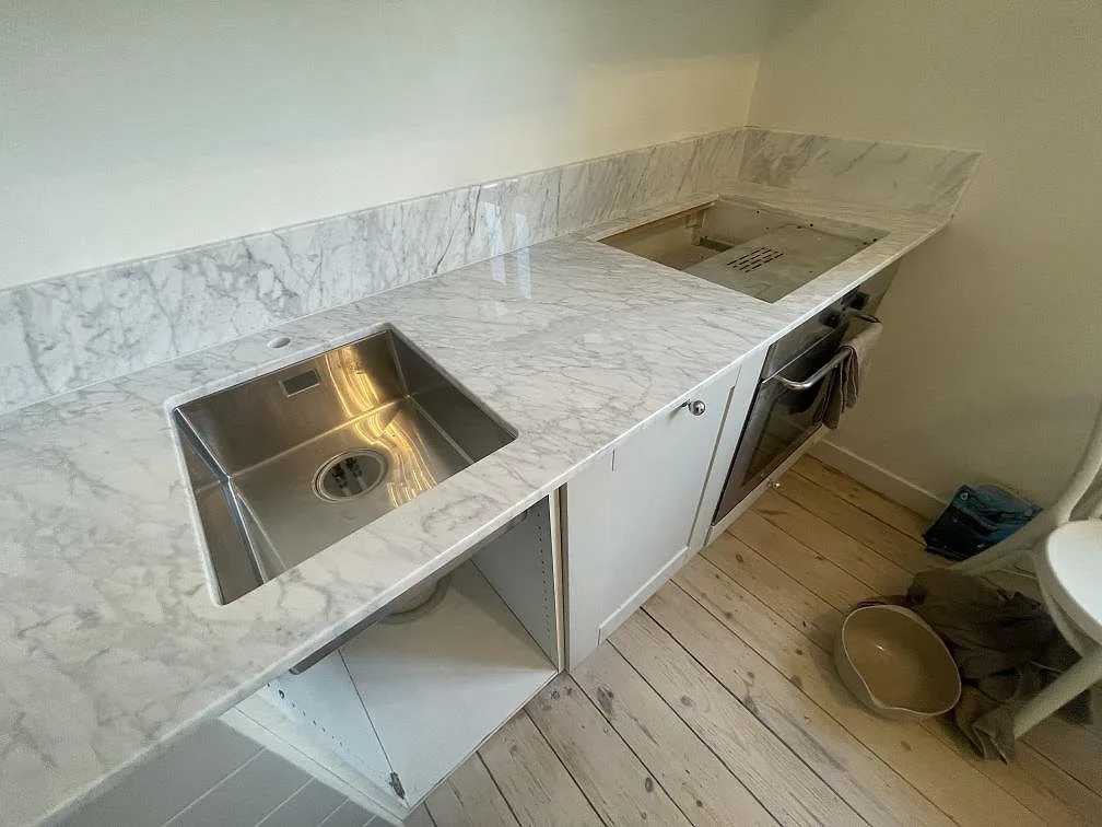 Närbild på polerad Carrara C marmorbänkskiva med underlimmad rostfri diskho och hällurtag ovanför ugnen i Stockholm.