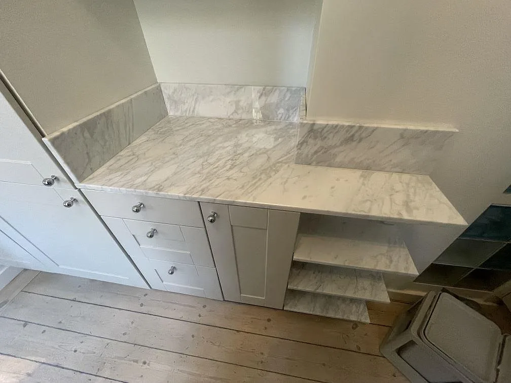 Bänkskiva i polerad Carrara C marmor med sidoretur, stänkskydd och öppna marmorklädda hyllplan i ett kök i Stockholm.