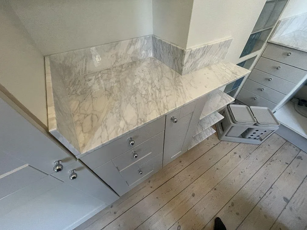 Carrara C marmorbänkskiva med grå ådring, uppvikt stänkskydd och hyllsektion under montering i Stockholm.