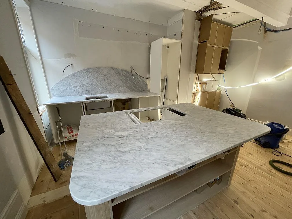 Bred vy över kök under montering med matt Carrara C marmor på köksö, bakre bänk och bågformat stänkskydd i Stockholm.