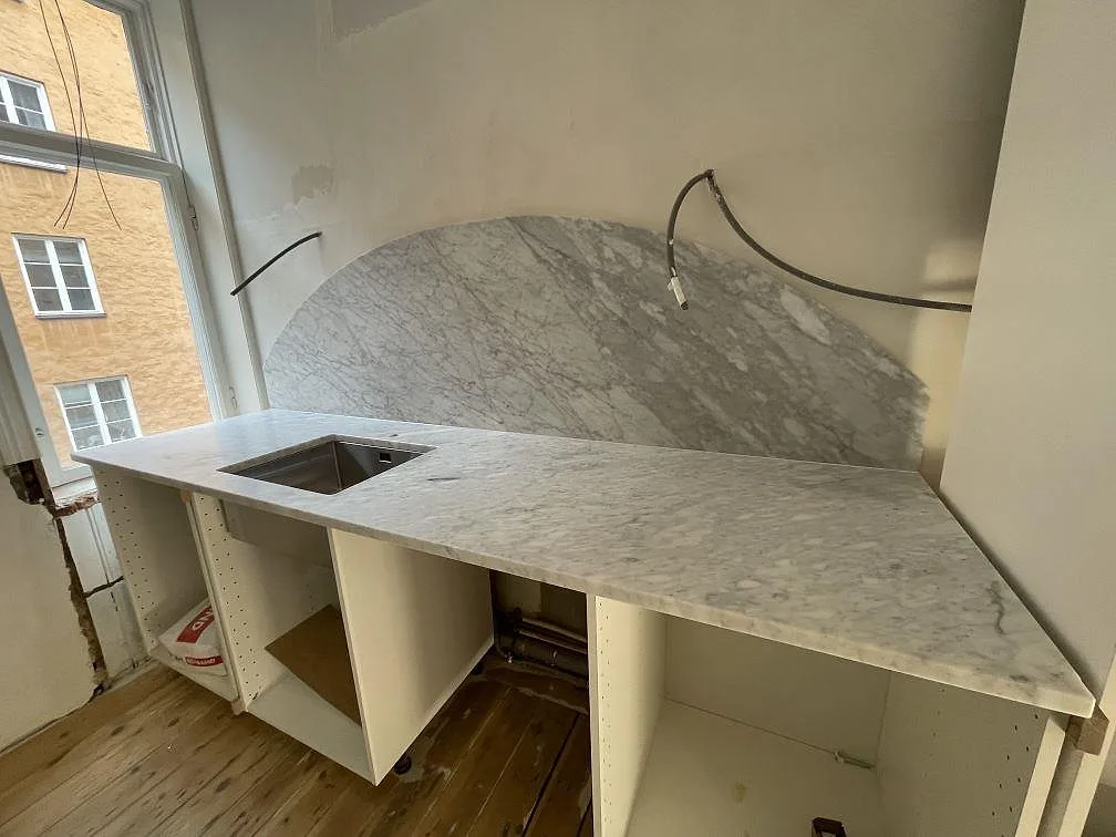 Matt Carrara C marmor som väggbänk med bågformat stänkskydd och öppna köksstommar vid fönster i Stockholm.