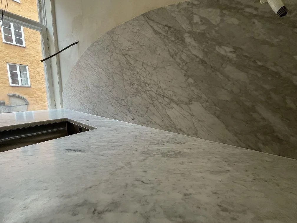 Detalj av matt Carrara C marmor med bågformat stänkskydd, diskzonens urtag och grå ådring i Stockholm.