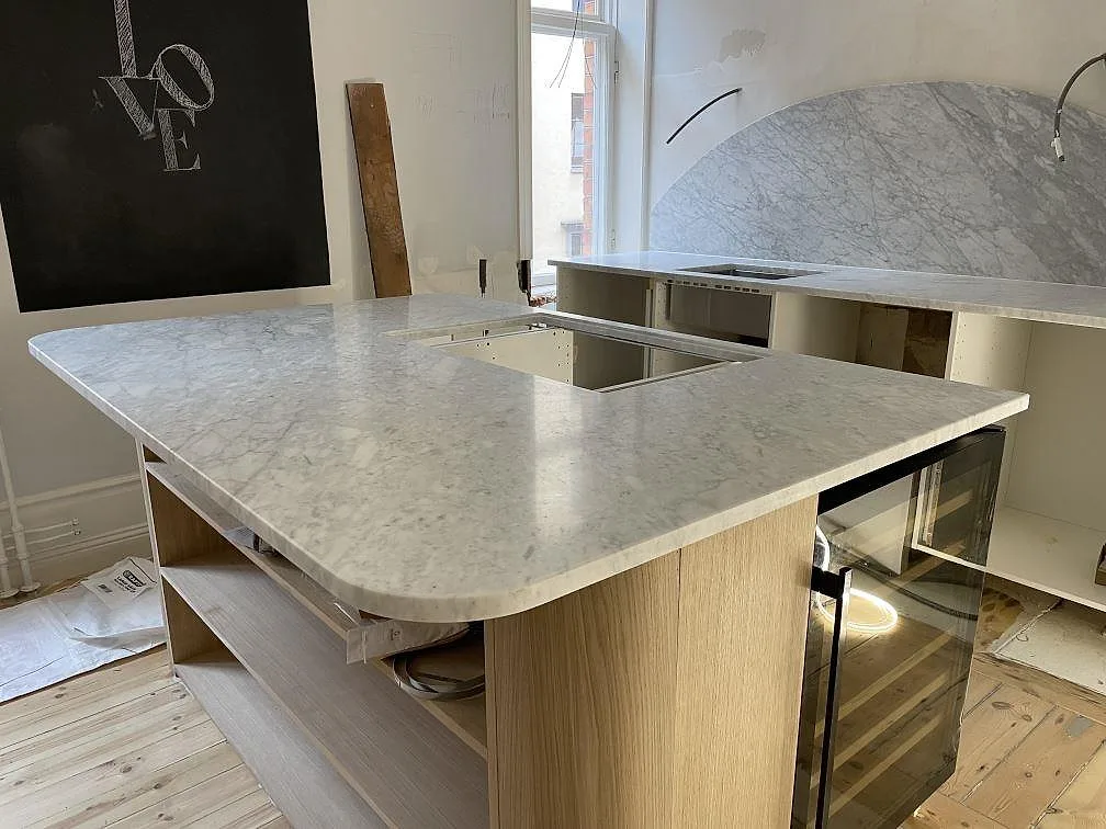 Närbild på köksö i matt Carrara C marmor med rundad kant, öppna stommar och synliga urtag i Stockholm.