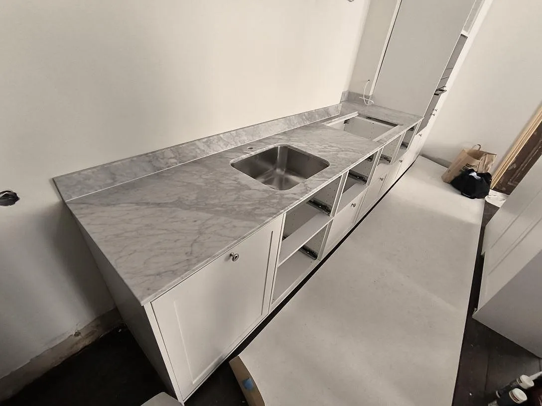 Köksbänkskiva i matt Carrara C marmor med rostfri diskho, hällurtag och uppvikt stänkkant i ett kök under montering i Stockholm.
