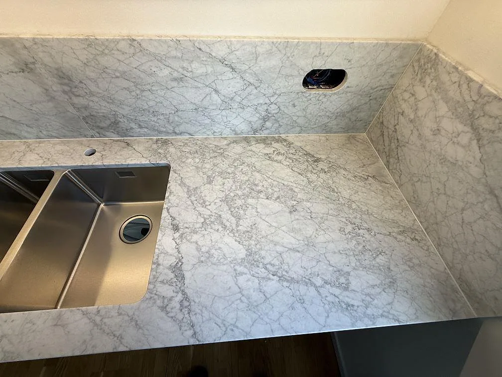 Detalj av matt Carrara C marmor vid rostfri diskho, vägguttag och sidostänkskydd i ett kök i Stockholm.