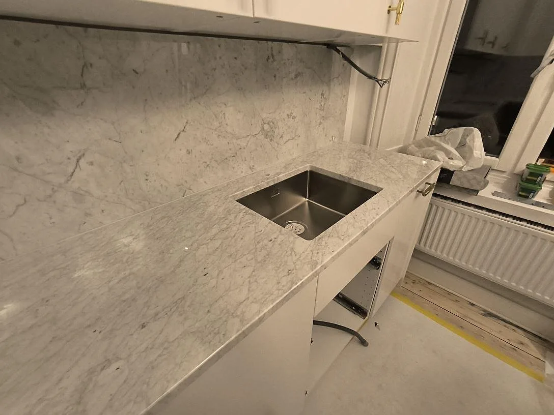 Detalj av polerad Carrara C marmor runt en monterad diskho med stänkskydd bakom i ett kök i Stockholm.
