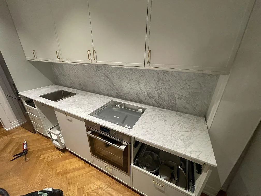 Matt köksbänkskiva i Carrara C marmor med lång bänkrad, stänkskydd och hällzon över ugn i Stockholm.