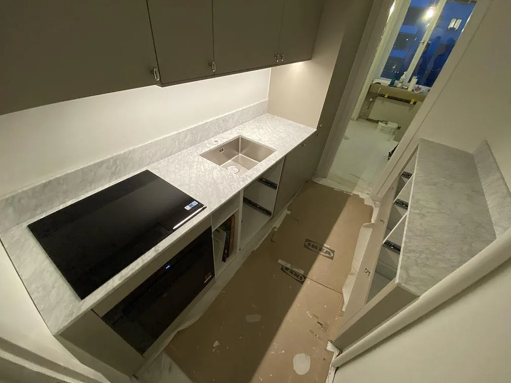 Höger bänkrad i matt Carrara C-marmor i ett kök i Stockholm med underlimmad rostfri diskho och svart induktionshäll infälld i stenen under taupefärgade överskåp.