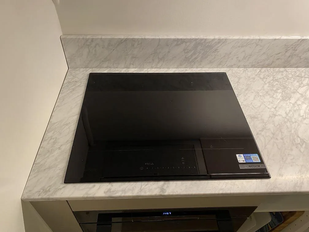 Svart Bosch induktionshäll infälld plant i en köksbänkskiva av matt Carrara C-marmor i Stockholm, med uppvikt stänkkant i samma marmor bakom.