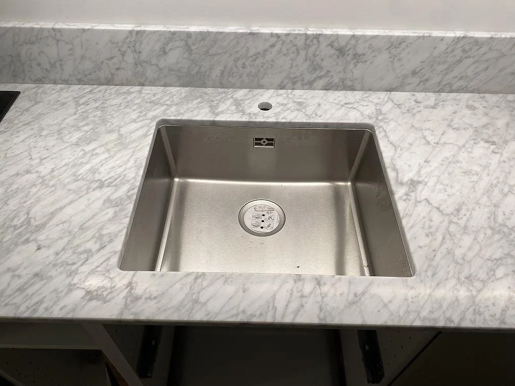 Underlimmad fyrkantig rostfri diskho i en köksbänkskiva av matt Carrara C-marmor i Stockholm, med fin grå ådring i den vita stenen.