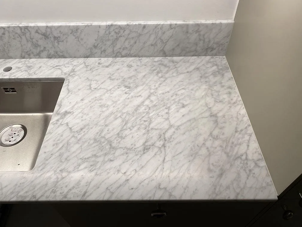 Top-down av matt Carrara C-marmor i ett kök i Stockholm med tät grå ådring och varma beige stråk, underlimmad rostfri diskho och uppvikt marmorstänkkant.