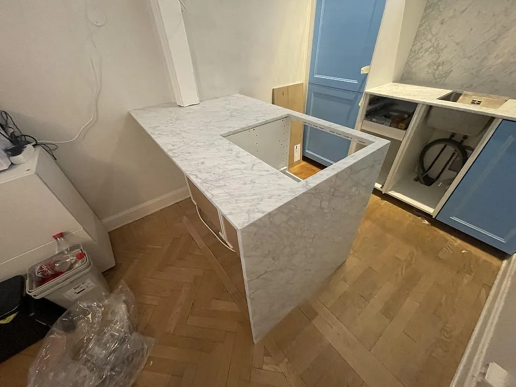 Halvö i Carrara C marmor sedd från sidan, med marmorklädd gavel ner mot fiskbensparketten och urtag för häll, i ett stockholmskök under montering.