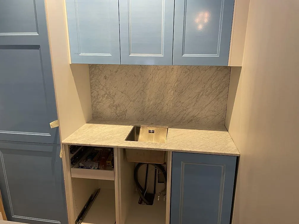 Underlimmad rostfri diskho i en bänkskiva av matt Carrara C marmor, med marmorstänkskydd ända upp till de ljusblå överskåpen i ett kök i Stockholm.