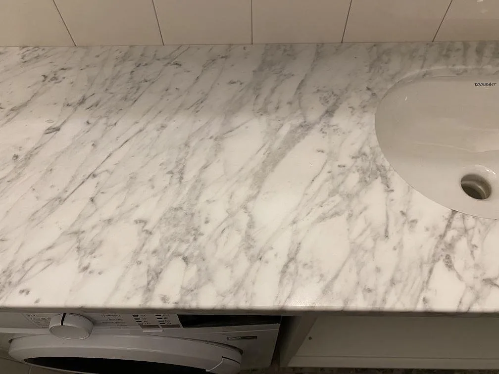 Bänkskiva i matt Carrara C-marmor sedd längs med i ett badrum i Stockholm, med mjuk grå ådring och det underlimmade Duravit-handfatet till höger.