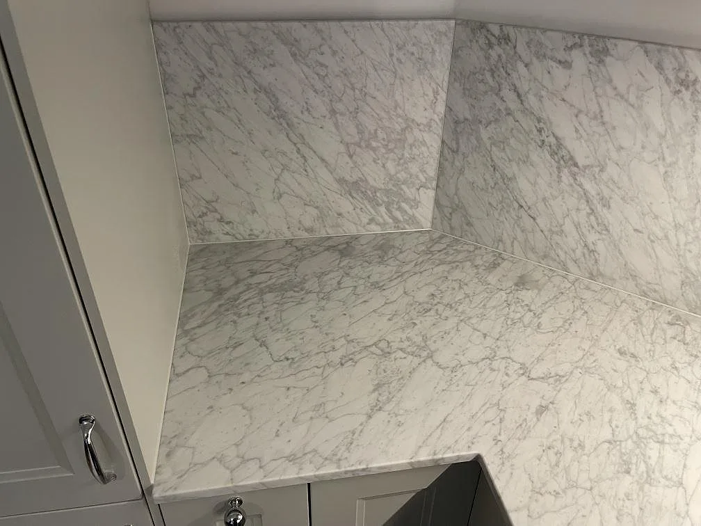 Hörndetalj där matt Carrara C bänkskiva möter en heltäckande marmorstänkvägg ovanför en ljusgrå skåkslucka i ett kök i Stockholm.