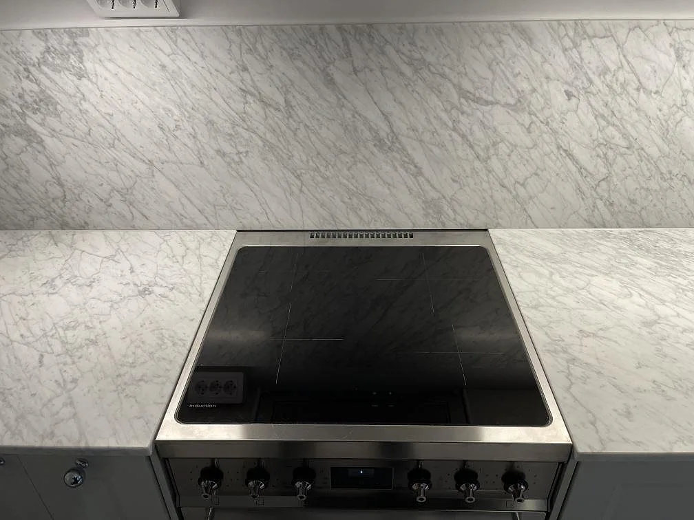Fristående induktionsspis infogad i bänkskiva av Carrara C marmor med matt finish, mot en heltäckande marmorstänkvägg i ett kök i Stockholm.