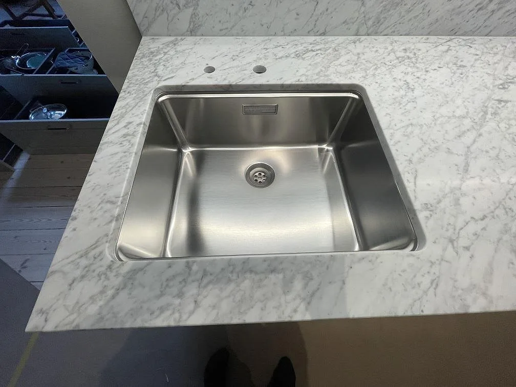 Närbild på underlimmad rostfri diskho med två blandarhål i en köksbänkskiva av matt Carrara C-marmor i Stockholm.