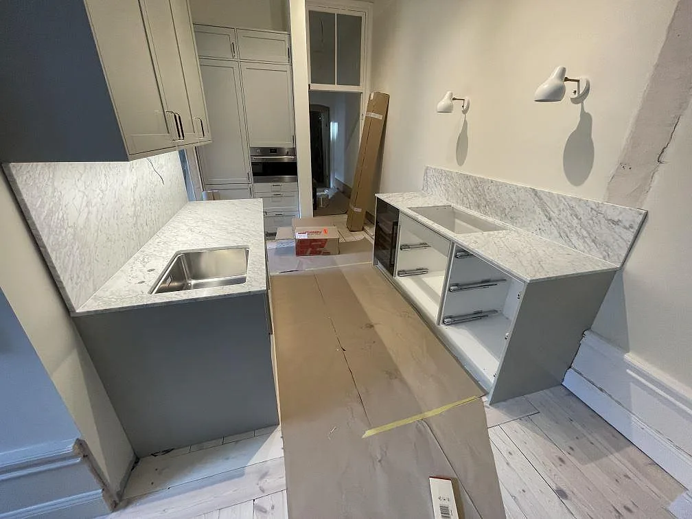 Vinkelkök under montering med två bänkar i matt Carrara C-marmor, marmorstänkväggar och delvis öppna lådstommar i en stockholmsvåning.