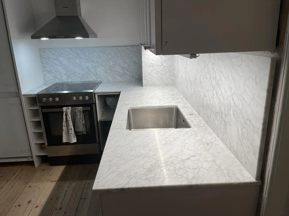 Vit Carrara C marmor i kök i Stockholm med fristående rostfri spis, fläktkåpa och underlimmad diskho i den matta bänkskivan.
